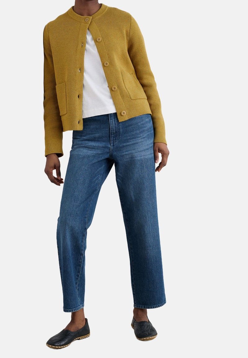 Personne portant un cardigan moutarde boutonné sur une chemise blanche, un jean bleu à jambes larges et des chaussures noires sans lacets, debout les jambes écartées.