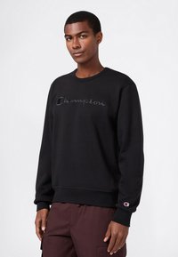 Giovane uomo in felpa nera Champion con logo sul petto e sulla manica, abbinata a pantaloni marrone scuro, in piedi contro uno sfondo semplice.