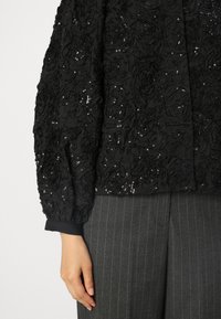 Schwarze Bluse mit strukturiertem Rosenmuster und Pailletten, mit Puffärmeln und schwarzem Bündchen. Kombiniert mit grauen schmal gestreiften Hosen.