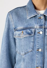 Giacca in denim blu chiaro con design classico, cuciture visibili, due tasche sul petto, bottoni argentati e superficie strutturata.