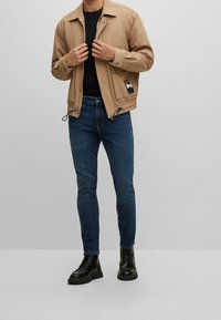 Hombre vistiendo una chaqueta beige, camisa negra, jeans azul oscuro y botas negras con cordones, de pie contra un fondo gris.