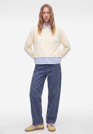Ung person står med hænderne bag ryggen iført en cremefarvet sweater over en blåstribet skjorte, blå bukser med nålestriber og beige sneakers.