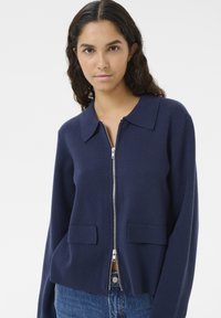 Marinblå zip-up cardigan i mjukt material, med klassisk krage, två framfickor och avslappnad passform.
