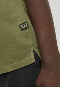 T-shirt in cotone verde oliva con spacco laterale, dotata di un'etichetta nera G-Star sul lato sinistro. Tessuto morbido, design aderente.