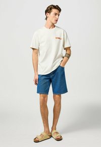 Camiseta corta de manga corta color crema con el logo "WRANGLER", combinada con pantalones cortos de denim azul y sandalias beige con una tira ancha y hebilla.