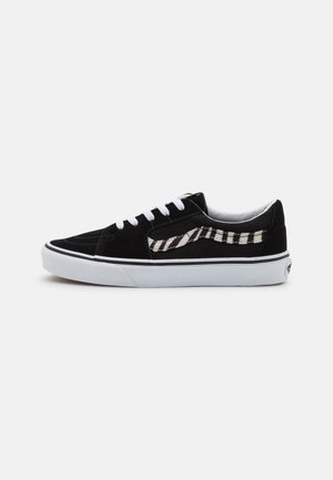 Zapatilla negra de caña baja con cordones blancos, suela de goma blanca y una franja lateral con patrón de cebra en blanco y negro.