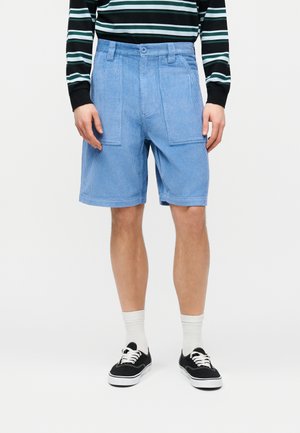 Személy világoskék kordbársony rövidnadrágban, fekete-fehér csíkos hosszú ujjú ingben, fehér zokniban és fekete fűzős tornacipőben.