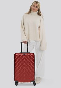 Mujer con un suéter beige y pantalones blancos de pie junto a una gran maleta roja rígida con un asa negra extendida.