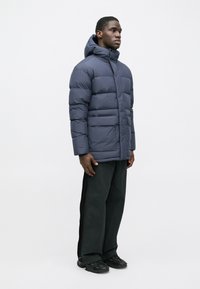 Ναυτικό πούφο μπουφάν με κουκούλα, διαθέτοντας σχεδίαση με ραφές, μπροστινές τσέπες και ευθεία γραμμή. Φορεμένο πάνω από σκούρες παντελόνες και μαύρα παπούτσια.