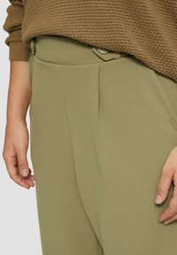 Olijfgroene broeken met een getailleerde pasvorm, voorzien van een knoopsluiting en voorplooien. De stof heeft een gladde textuur.