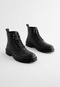 Botas de tobillo negras con cordones, hechas de cuero con un acabado texturizado, que presentan una suela gruesa y una lengüeta en la parte posterior para facilitar su puesta.