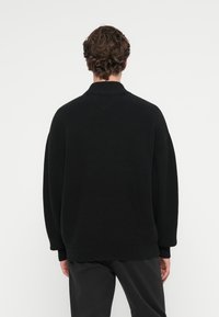 Μαύρο πλεκτό πουλόβερ με ψηλό γιακά, με χαλαρή εφαρμογή και ριγέ υφή. Οι μανσέτες και το τελείωμα είναι επίσης ριγέ.