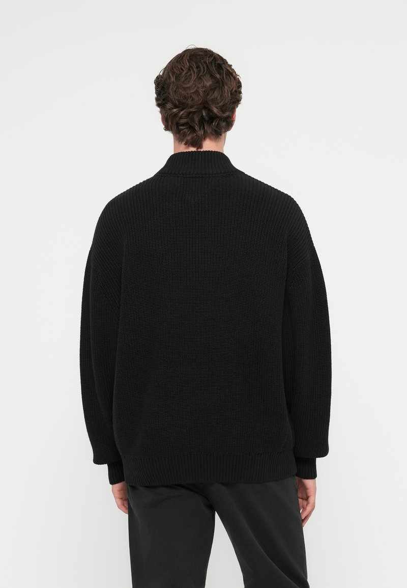Zwarte gebreide trui met een hoge kraag, in een losse pasvorm en geribbeld textuur. De manchetten van de mouwen en de zoom zijn ook geribbeld.