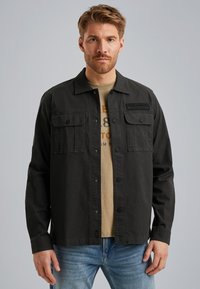 Veste de travail en coton noir avec deux poches poitrine, boutons-pression et un patch logo. Portée sur un T-shirt marron clair et un jean bleu.