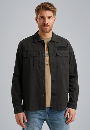 Chaqueta de trabajo de algodón negra con dos bolsillos en el pecho, botones a presión y un parche de logo. Lleva puesta una camiseta color marrón claro y unos vaqueros azules.
