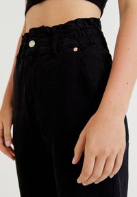 Svarta byxor med elastisk midja, volang i midjan, sidofickor och metallaccenter, som visar en slät, strukturerad tyg.