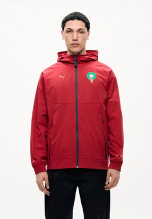 Homme portant une veste zippée rouge avec un emblème vert et or sur la poitrine et un pantalon noir, debout devant un fond blanc.