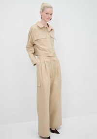 Tuta utility beige di grandi dimensioni con due ampi taschini sul petto, collo a camicia e design a gamba larga. Tessuto testurizzato, dettagli plissettati.