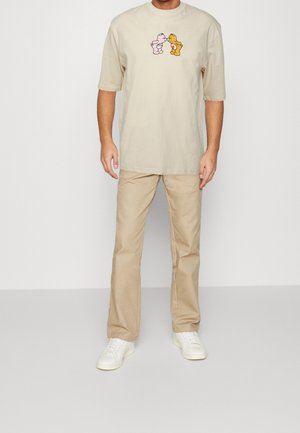 Beige Baumwoll-T-Shirt mit einem verspielten pinken und gelben Bärendruck, kombiniert mit hellbeigen Hosen und weißen Sneakern.