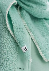 Écharpe vert menthe avec tissu pelucheux et texturé, finition lisse. Présente un bord blanc cousu et une petite étiquette avec un logo.