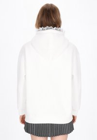 Felpa bianca oversize con bordo in pizzo strutturato sul cappuccio, abbinata a una gonna grigia plissettata. Il tessuto appare morbido e liscio.