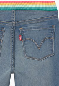 Modré denimové džínsy s vícerozměrným žebrovaným pasem, obsahující dvě zadní kapsy a charakteristické švy ve tvaru V. Viditelný červený štítek značky.