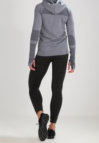 Personne portant un haut à manches longues à capuche gris et des leggings noirs, se tenant avec un pied légèrement levé, devant un fond uni.