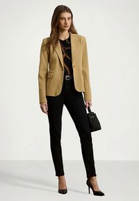 Γυναίκα που φοράει ένα καμηλί μπλέιζερ, patterned μαύρη μπλούζα, μαύρο παντελόνι και μαύρες γόβες, κρατώντας μια μικρή μαύρη τσάντα με λεπτομέρεια δαχτυλιδιού.