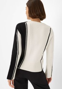 Femme portant un pull en maille torsadée noir et blanc avec des rayures verticales, vue de dos, associé à un pantalon noir.