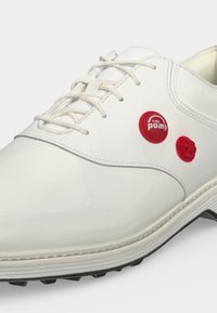 Chaussure de sport en cuir blanc avec des lacets blancs et deux boutons-pompes rouges circulaires sur le côté au-dessus de la semelle.