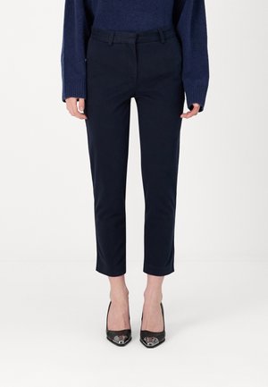 Vrouw die marineblauwe cropped broek, marineblauwe trui en zwarte pumps met puntige neus draagt, staand tegen een witte achtergrond.
