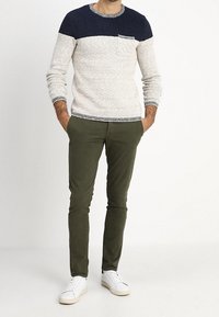 Uomo che indossa un maglione lavorato a maglia blu navy e beige, pantaloni slim verde oliva e sneakers bianche, in piedi con le mani in tasca su uno sfondo bianco.
