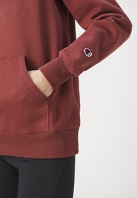 Burgundi pulóver kenguruzsebbelt, bordás mandzsettákkal és aljjal, melyen egy kis hímzett logó található az ujjon. Puha szövettextúra.