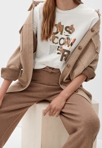 Beige oversized wool coat with buttons, white t-shirt featuring colorful graphic text, and matching beige knit pants with a drawstring.