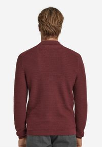 Burgundy stickad tröja med ett texturerat mönster, ribbade ärmslut och krage. Bakre vy framhäver det vävda mönstret och den enfärgade designen.