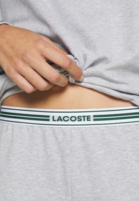 Grijze katoenen top met een losse pasvorm, hand die de zoom vasthoudt. Groene en witte gestreepte elastische band met het etiket "LACOSTE" zichtbaar op de tailleband.