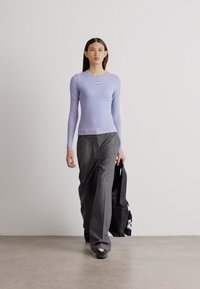 Hellblau-langes Oberteil mit subtiler Textur, kombiniert mit dunkel gestreiften, weit geschnittenen Hosen. Modell hält eine schwarze Tote-Bag.