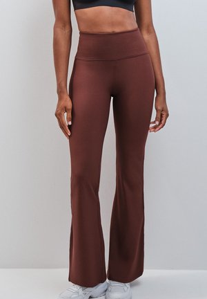 Femme portant un pantalon évasé marron taille haute et des baskets blanches, debout devant un fond gris clair.