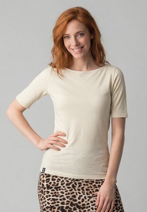 Young woman with red hair smiles, wearing a beige short-sleeve top and leopard print skirt, standing against a plain gray background.