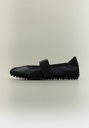 Zapato plano negro con suela texturizada y una sola tira ancha en la parte superior, mostrado de perfil sobre un fondo claro y sencillo.
