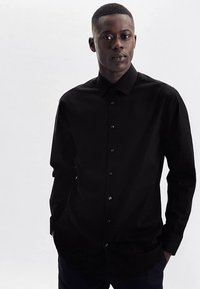 Schwarzes Hemd mit Knopfleiste aus glattem Stoff, mit spitzem Kragen, langen Ärmeln und dezenten Knopfdetails. In entspannter Pose getragen.