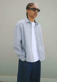 Camicia a maniche lunghe a righe blu chiaro, realizzata in tessuto morbido, indossata sopra una t-shirt bianca, abbinata a jeans larghi blu scuro e occhiali da sole scuri.