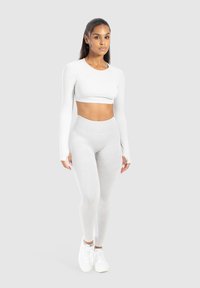 Jonge vrouw staat met een neutrale uitdrukking, gekleed in een witte lange mouw crop top, lichtgrijze high-waisted leggings en witte sneakers.