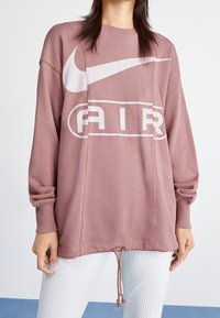Volný růžový svetr s velkým bílým logem Nike a grafikou "AIR". Obsahuje žebrované manžety a stahovací lem pro nastavitelnost.