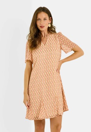 Femme aux longs cheveux bruns portant une robe courte à manches courtes, de couleur pêche et orange à motifs, longueur genou, debout avec une main sur la hanche devant un fond uni.
