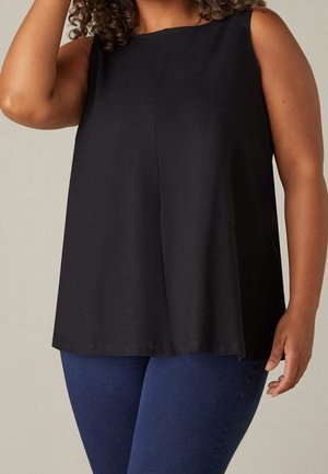 Donna che indossa un top nero senza maniche e jeans blu, con un braccio alzato che tocca i capelli e l'altro rilassato lungo il corpo.