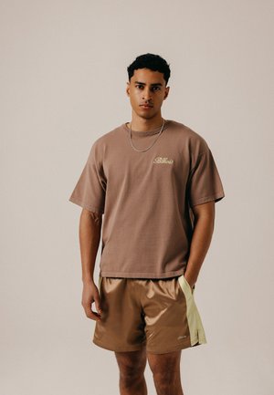 Ung mand står med den ene hånd i lommen, iført en brun oversized T-shirt og skinnende tofarvede shorts mod en ensfarvet baggrund.