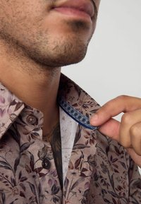 Camicia marrone con motivo floreale, dotata di un colletto con bottoni e un accento blu a contrasto sulla fodera interna del colletto.