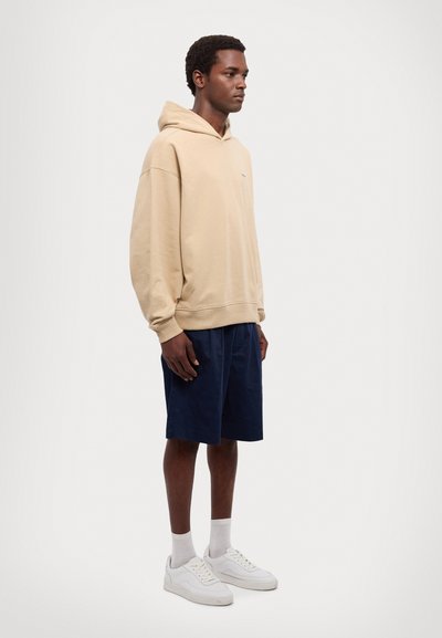 Hoodie bej, potongan santai, bahan lembut, kantong kanguru, dipasangkan dengan celana pendek navy, sneakers putih, kaus kaki polos, berdiri di latar belakang netral.