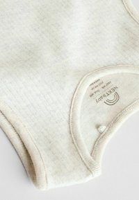 Light beige ribbed baby bodysuit with a rounded neckline, soft texture, and a snap closure at the bottom; includes brand labeling inside.